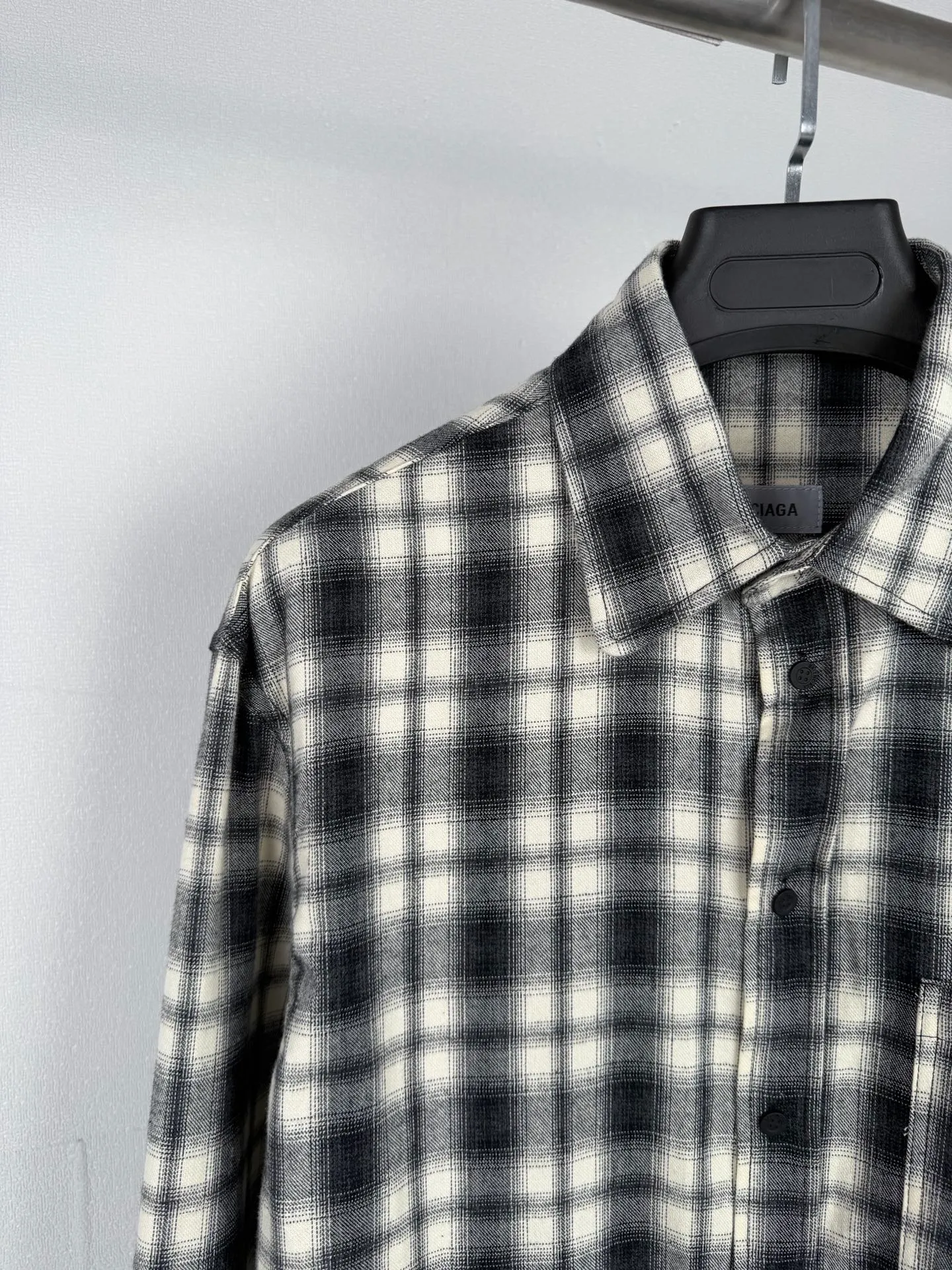 BALENCIAGA/balenciaga Black White plaid pocket embroidered logo shirt jacket The technical difficulties of this plaid shirt focus on fabric finishing, plaid matching and component sewing details. The fabric has been sanded and distressed to create a fine velvet feel on the surface, changing the original luster of the fabric and making the Black and White plaid colors transition softly. At the same time, the drape properties of the fabric itself are retained, taking into account the looseness of the upper body and the silhouette of the body, without appearing soft and shapeless. The biggest test of plaid garments comes from the alignment of the pieces. The front and rear body, large and small sleeves, and chest pockets are all independent pieces. In the cutting stage, the horizontal and vertical axes of the plaid need to be aligned, and the external chest bag is cut separately. The plaid on the bag surface needs to form a visual coordination with the main body of the garment. Cutting and typesetting will produce additional fabric loss, and there are high requirements for the cutting accuracy of the cutting bed typesetting and workers. The collar adopts a double-layer combined structure, and the collar surface and collar seat are double-stitched with even and regular stitching spacing to ensure the lapel's long-lasting shape and resistance to warping and deformation after repeated bending. The placket is sewn with regular topstitching and paired with Black matte buttons. The button points need to be aligned with the checkered lines to avoid damaging the overall visual order. The splicing of the cuffs also takes into account the grain direction, and the overlocking process reinforces the seams to improve the durability of the garment. After the entire garment is sewn, it also undergoes a garment washing process to further strengthen the Retro texture of the fabric, unify the tone and texture of the entire garment, weaken the harshness of industrial production, and finally present a relaxed Retro garment effect. Size: 46 48 50 52